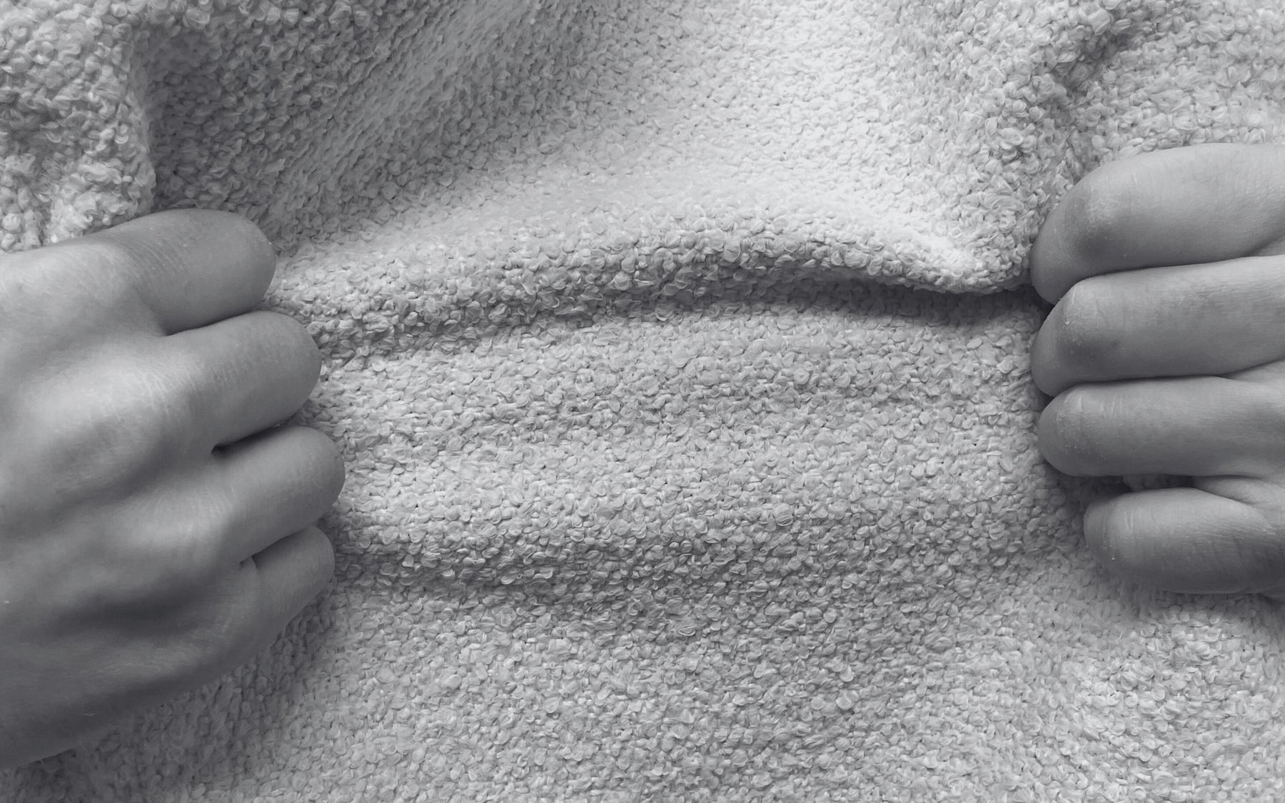 A close-up black and white image of two hands pulling at a textured upholstery fabric, testing its elasticity and durability.