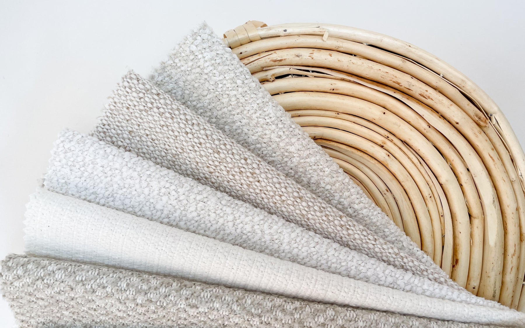 A collection of textured upholstery fabrics in varying shades of white and light gray, neatly folded and stacked in ascending order of texture complexity, displayed against a natural, curved rattan chair back.